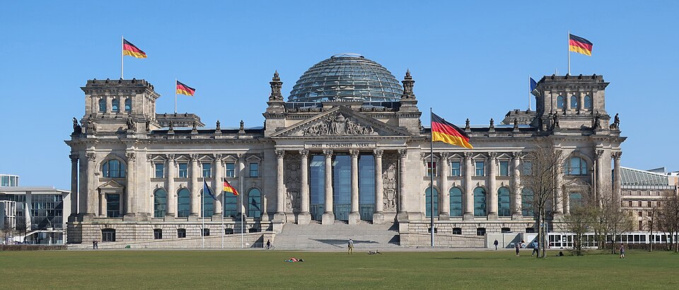 Reichstag_Berlin - EXPOsed Berlin's Reichstag building in bright sun, featuring its glass dome and flags flying on a clear day, with a green lawn in front.
