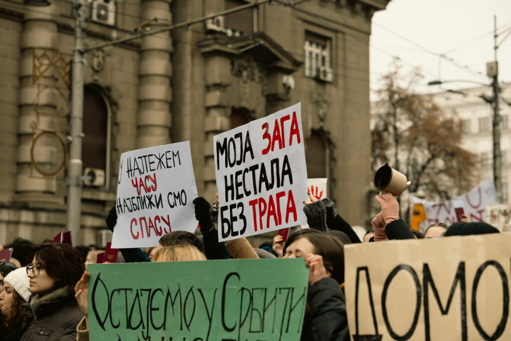 Crowd of protesters on a city street, holding white signs with Cyrillic text; a person holds a megaphone on the right.