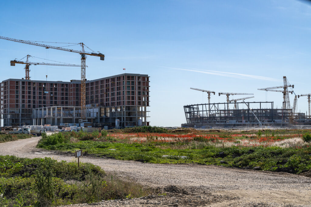 EXPO stadium construction - EXPOsed Construction site with multiple tall cranes over a partially built brick building under a clear blue sky, with grassy foreground and dirt path.