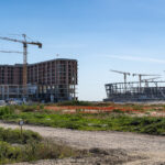 Construction site with multiple tall cranes over a partially built brick building under a clear blue sky, with grassy foreground and dirt path.